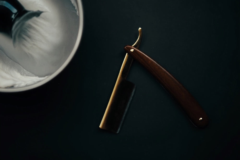 Straight razor, lather bowl, and brush laid out on a dark surface